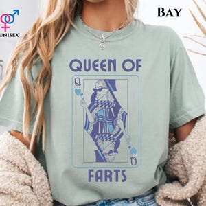 May include: A light green unisex t-shirt with the text "QUEEN OF FARTS" in blue. The shirt features a playing card design with a woman wearing sunglasses. The word "BAY" is printed at the top right.