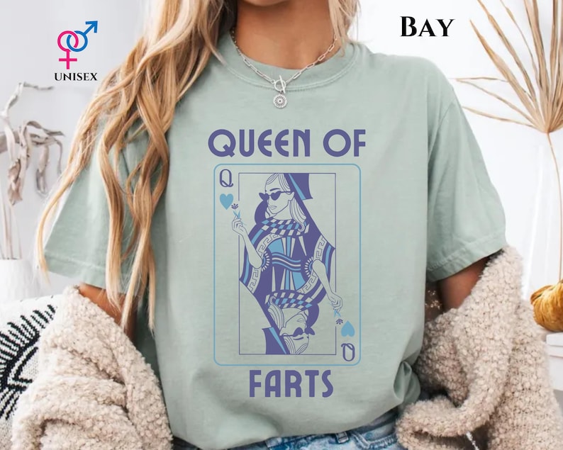 May include: A light green unisex t-shirt with the text "QUEEN OF FARTS" in blue. The shirt features a playing card design with a woman wearing sunglasses. The word "BAY" is printed at the top right.
