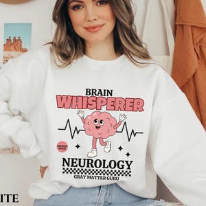May include: White sweatshirt with a graphic of a brain with a smiling face and the text "Brain Whisperer" and "Neurology Gray Matter Guru".