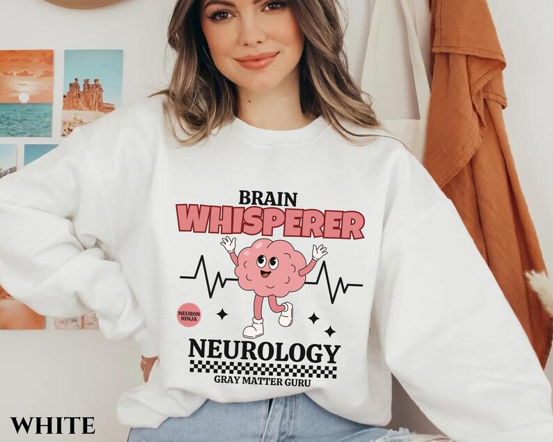 May include: White sweatshirt with a graphic of a brain with a smiling face and the text "Brain Whisperer" and "Neurology Gray Matter Guru".