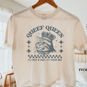 May include: An ivory short-sleeve t-shirt featuring a dark blue graphic of a cat wearing a crown. The text reads "QUEEF QUEEN" above the cat and "IT'S NOT A FART, IT'S PUSSY ART" below, with a checkered pattern and star accents surrounding the design.