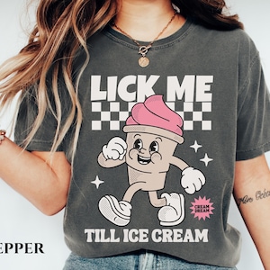 May include: A gray t-shirt with a cartoon ice cream cone graphic and the text "Lick Me Till Ice Cream". The ice cream cone is pink and white with a smiling face. The text "Cream Dream" is in a small circle on the bottom right corner of the shirt.