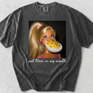 May include: A charcoal gray short-sleeve t-shirt featuring a graphic of a blonde doll with a high ponytail holding a deviled egg to its mouth. Below the graphic, the white cursive text reads, "put them in my mouth." The shirt has a vintage wash.