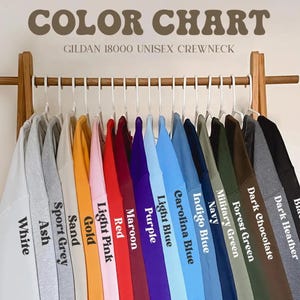 May include: Color chart showing 15 different colors of Gildan 18000 unisex crewneck sweatshirts. The colors are: White, Ash, Sport Grey, Sand, Gold, Light Pink, Red, Maroon, Purple, Light Blue, Carolina Blue, Indigo Blue, Navy, Military Green, Forest Green, Dark Chocolate, Dark Heather, and Black.