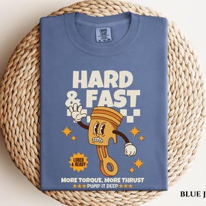 Puede incluir: Camiseta de mezclilla azul con una ilustraci&oacute;n de dibujos animados de un pist&oacute;n con el texto "Hard & Fast" y "More Torque, More Thrust - Pump It Deep" impreso en la parte delantera.