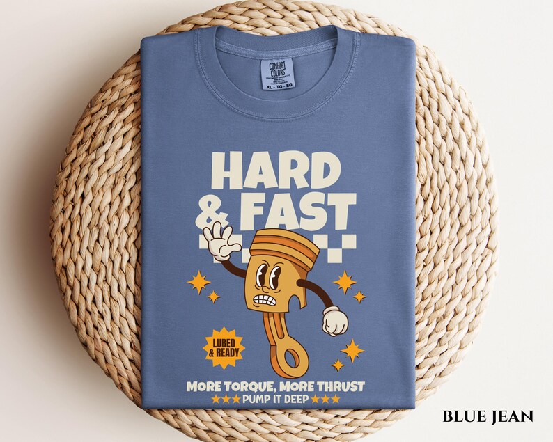 Puede incluir: Camiseta de mezclilla azul con una ilustraci&oacute;n de dibujos animados de un pist&oacute;n con el texto "Hard & Fast" y "More Torque, More Thrust - Pump It Deep" impreso en la parte delantera.