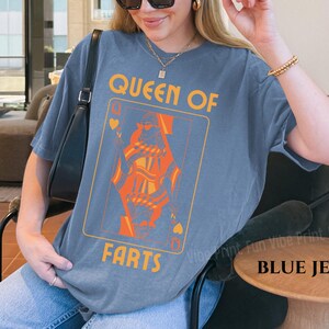 May include: Blue jean-colored t-shirt featuring a playing card design with the text "QUEEN OF FARTS" in orange and yellow. The graphic depicts a queen card. The shirt is a relaxed fit.