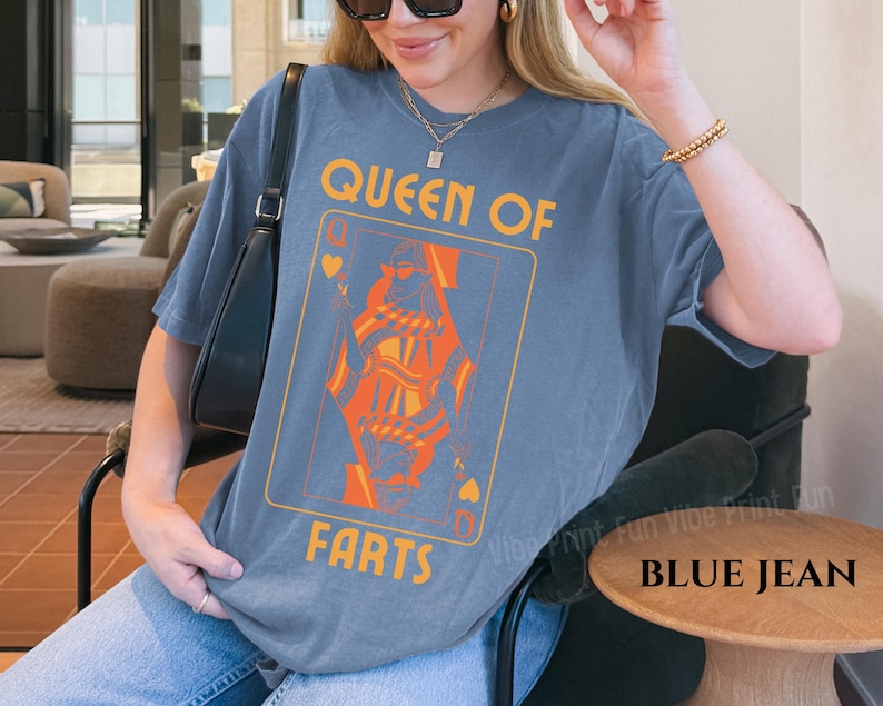 May include: Blue jean-colored t-shirt featuring a playing card design with the text "QUEEN OF FARTS" in orange and yellow. The graphic depicts a queen card. The shirt is a relaxed fit.