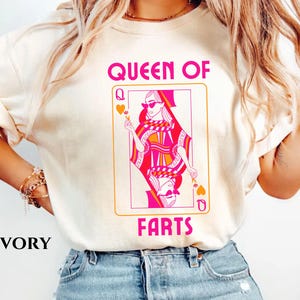 May include: Ivory-colored t-shirt featuring a playing card design with a woman wearing sunglasses. The card reads "QUEEN OF FARTS" in pink lettering. The card design is in shades of pink, orange, and white.