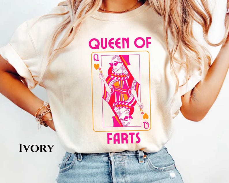 May include: Ivory-colored t-shirt featuring a playing card design with a woman wearing sunglasses. The card reads "QUEEN OF FARTS" in pink lettering. The card design is in shades of pink, orange, and white.