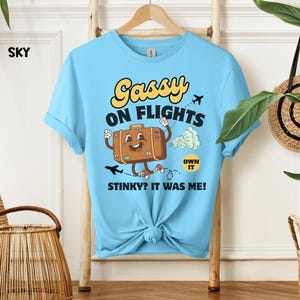 May include: Sky blue t-shirt with a cartoon graphic of a suitcase with arms and legs, the text "Sassy on Flights" and "Stinky? It was me!" in yellow and black. The shirt is knotted at the bottom.