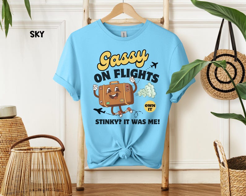May include: Sky blue t-shirt with a cartoon graphic of a suitcase with arms and legs, the text "Sassy on Flights" and "Stinky? It was me!" in yellow and black. The shirt is knotted at the bottom.