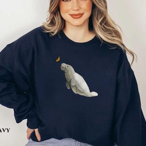 May include: A navy blue sweatshirt with a graphic of a manatee and a butterfly. The sweatshirt has a crew neck and long sleeves.