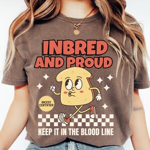 May include: A brown t-shirt with the text "INBRED AND PROUD" in red and orange letters. A cartoon bread character with a face and legs is walking on a checkered pattern with the text "KEEP IT IN THE BLOOD LINE". A small red circle with the text "INCEST CERTIFIED" is in the upper left corner.