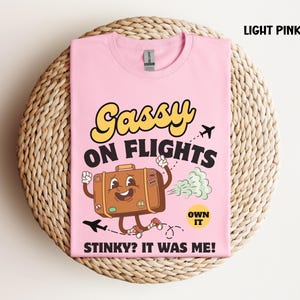 May include: Light pink t-shirt with the text "Gassy on Flights" in yellow and black. A cartoon suitcase with arms and legs is running, emitting a green cloud. The text "Stinky? It was me!" is below.