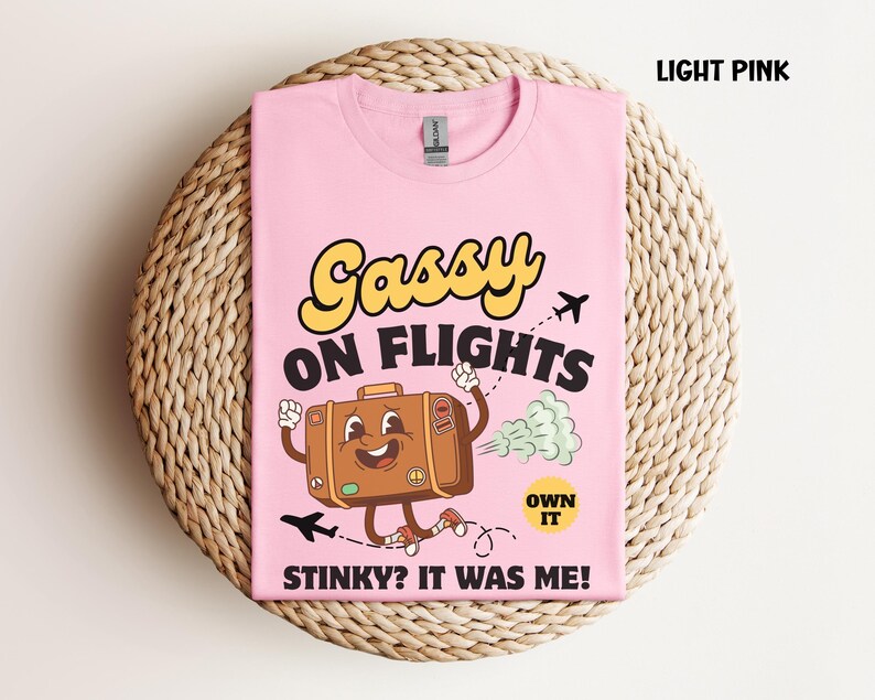 May include: Light pink t-shirt with the text "Gassy on Flights" in yellow and black. A cartoon suitcase with arms and legs is running, emitting a green cloud. The text "Stinky? It was me!" is below.