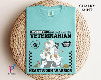 May include: A light blue unisex t-shirt with the text "POWERED BY: CAT HAIR & COFFEE VETERINARIAN HEARTWORM WARRIOR". The shirt features a graphic of various animals, including cats, dogs, a bird, and a turtle. The shirt is labeled "CHALKY MINT".