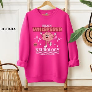 May include: A pink sweatshirt with a brain graphic and the text "Brain Whisperer Neurology Gray Matter Guru".