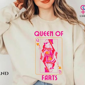 May include: Sand-colored unisex sweatshirt with the text "QUEEN OF FARTS" above a playing card graphic. The card features a stylized queen in pink and orange, holding a heart. The word "UNISEX" is printed on the upper right.