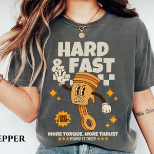 Puede incluir: Una camiseta gris oscuro con una ilustraci&oacute;n de dibujos animados de un pist&oacute;n con una cara. El texto "HARD & FAST" est&aacute; por encima del pist&oacute;n y "MORE TORQUE, MORE THRUST ★★★PUMP IT DEEP ★★★" est&aacute; debajo del pist&oacute;n. El texto "LUBED & READY" est&aacute; en un peque&ntilde;o rect&aacute;ngulo por encima del pist&oacute;n.
