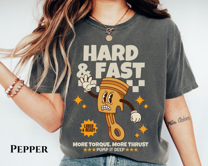 Puede incluir: Una camiseta gris oscuro con una ilustraci&oacute;n de dibujos animados de un pist&oacute;n con una cara. El texto "HARD & FAST" est&aacute; por encima del pist&oacute;n y "MORE TORQUE, MORE THRUST ★★★PUMP IT DEEP ★★★" est&aacute; debajo del pist&oacute;n. El texto "LUBED & READY" est&aacute; en un peque&ntilde;o rect&aacute;ngulo por encima del pist&oacute;n.