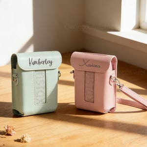 May include: Two personalized pastel-colored camera cases. One is mint green with the name "Kimberley" and the other is pink with the name "Xaviera". Both cases have a strap and a rectangular window.