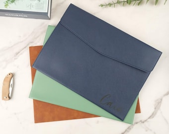 May include: Three leather laptop sleeves stacked on top of each other. The top sleeve is navy blue, the middle sleeve is light green, and the bottom sleeve is brown. The navy blue sleeve has the name "Carrie" engraved on it.
