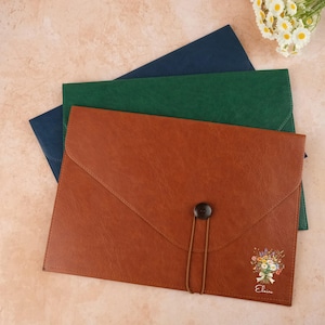 May include: Three leather document folders in brown, green, and navy blue. The brown folder has a button closure and a floral design with the name "Elaine" printed on it. A bouquet of white daisies is in the upper right corner.