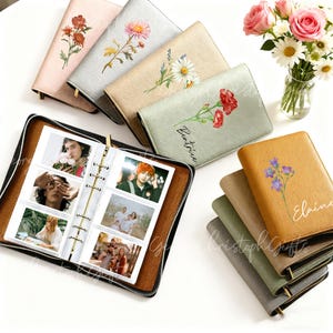 May include: A collection of personalized photo albums in various colors, including pink, silver, and tan, each adorned with floral designs and names. One album is open, displaying photos. A vase of flowers sits nearby.