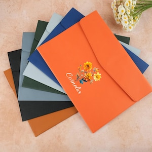 May include: A collection of colorful document folders in various shades, including orange, blue, and black. The orange folder features a floral design and the name "Cornelia". The folders are arranged in a fan shape, showcasing their different colors and textures.