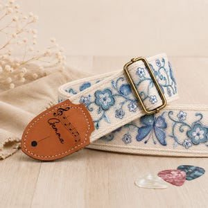 May include: A cream-coloured guitar strap featuring blue floral and butterfly embroidery. It includes a tan leather end tab with the name Gemma and a musical staff printed on it, along with a brass-toned metal adjustment buckle and three guitar picks.