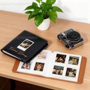 May include: A black leather travel diary with the words "Travel Diary" in gold script, next to a photo. The diary is open, revealing several photos. A black instant camera sits nearby. A small potted plant is in the background.