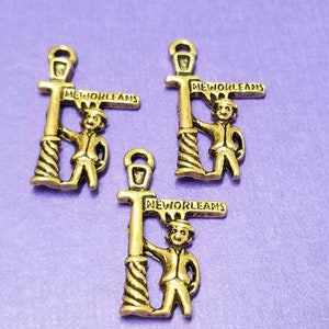 May include: Three gold-tone metal charms featuring a cartoon figure standing next to a street lamp with a sign that reads "NEW ORLEANS".