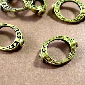 May include: Close-up of several antique brass-colored oval connector rings. Each ring features a decorative design of small circles and lines around the outer edge. The connectors have a small cylindrical element on each end.
