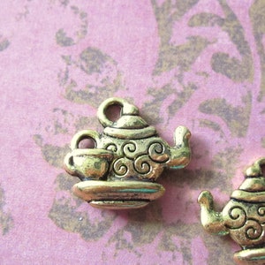 May include: Two gold-tone metal tea pot charms with a swirl design. Each charm features a teapot with a cup and saucer.