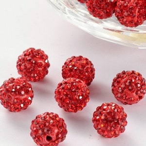 May include: Red rhinestone beads, a close-up of a group of small red beads covered in rhinestones. The beads are scattered on a white surface.
