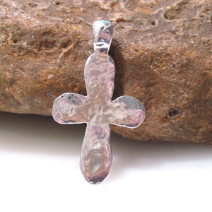May include: A silver cross pendant with a hammered texture. The pendant is displayed on a brown rock.
