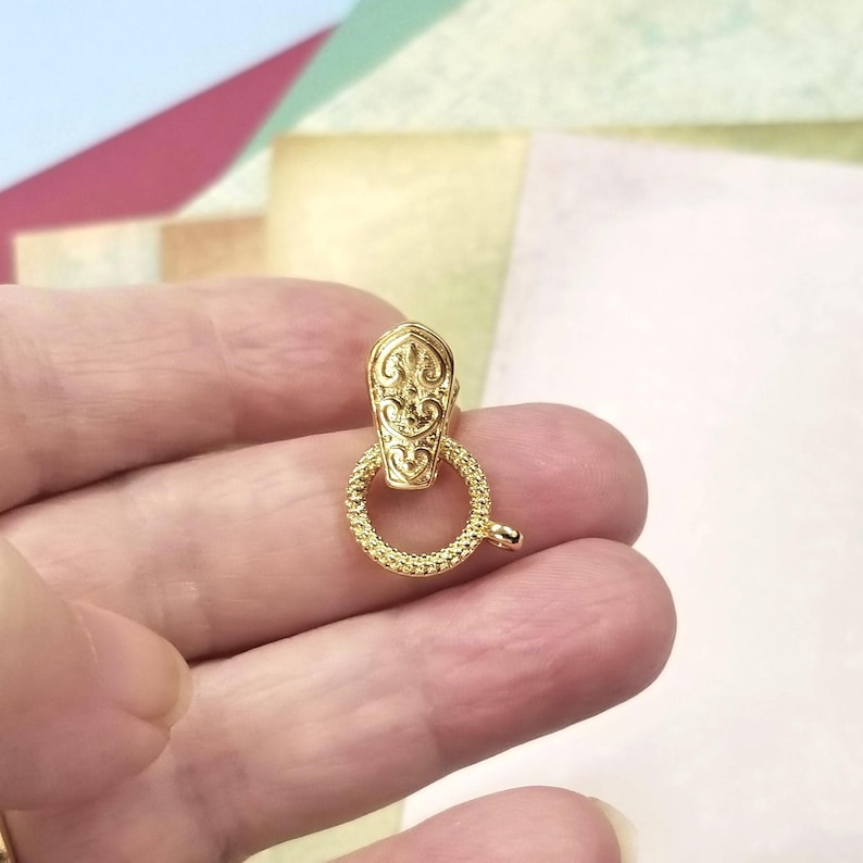 May include: A gold-colored clasp with an ornate design. The clasp features a decorative top piece and a textured ring with a small lever. The clasp is designed for jewelry making and crafting.