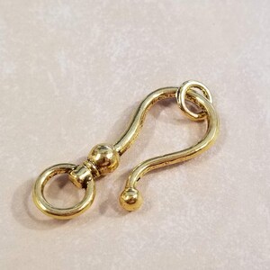 May include: Gold-toned metal hook with a round loop at one end and a curved hook at the other end. The hook is designed to attach to a chain or other jewelry.