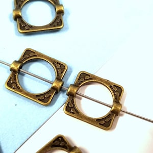 May include: Antique brass square-shaped metal connector with a circular opening in the center. The connector has a decorative design around the opening and is strung on a thin wire.