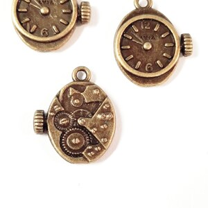May include: Three antique gold-toned metal watch charms. One charm features a clock face with Roman numeral markings, another charm features a clock face with Arabic numeral markings, and the third charm features a detailed clockwork mechanism.