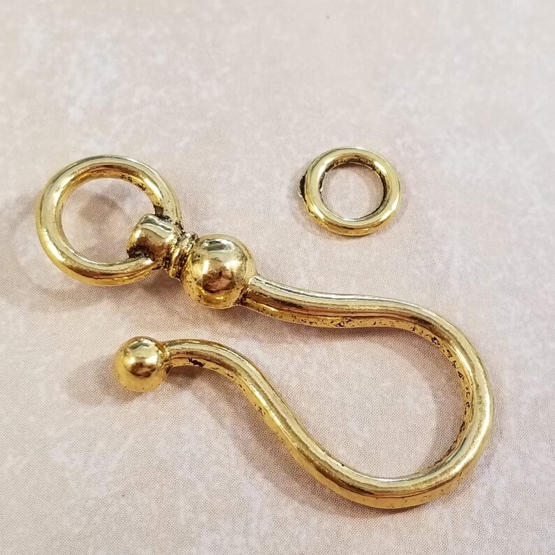May include: A gold-toned metal swivel clasp with a round loop and a decorative ball. The clasp is designed to attach to a chain or other jewelry.