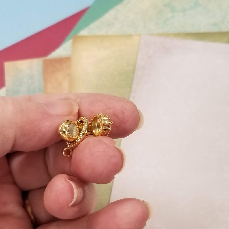May include: A small, gold-colored jewelry clasp with a round, iridescent stone and a decorative, textured design. The clasp is held between fingers against a colorful paper background.