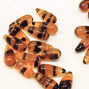 May include: A collection of amber glass teardrop beads with black and brown stripes. The beads are arranged on a white surface.