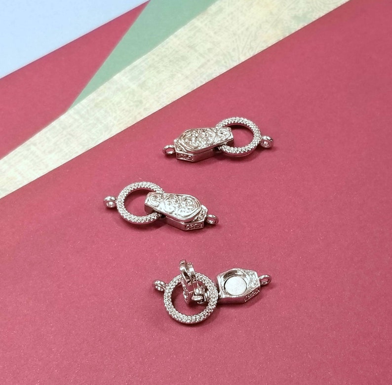 May include: Three silver-toned metal clasps with a round ring and a decorative design. The clasps are on a red surface.
