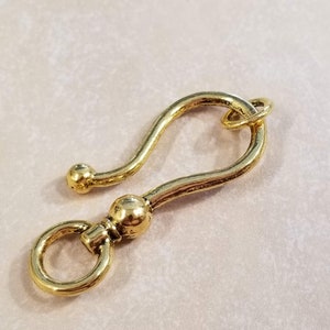 May include: A gold-toned metal hook with a rounded end and a small loop at the top. The hook is designed to be attached to a chain or other jewelry.