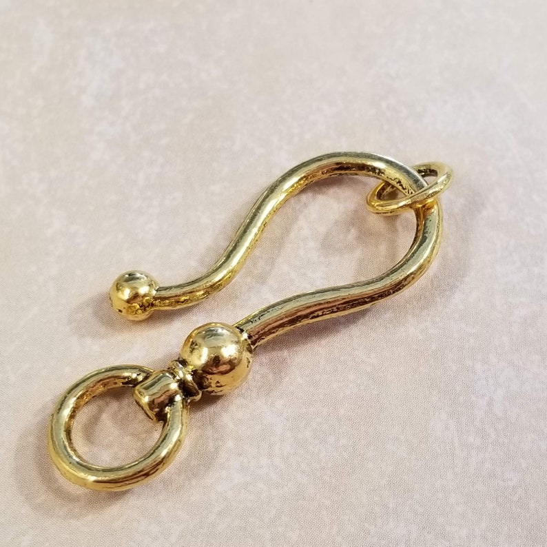 May include: A gold-toned metal hook with a rounded end and a small loop at the top. The hook is designed to be attached to a chain or other jewelry.