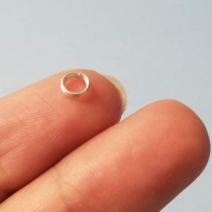 May include: A small, open, silver-colored jump ring, resting on a fingertip. The ring is circular with a small gap, suggesting it is designed to be opened and closed. The background is a soft, light blue.