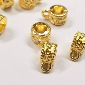 May include: Gold-toned metal pendant bails with a decorative design. The bails have a loop at the top for attaching a pendant and a small loop at the bottom for attaching a chain.