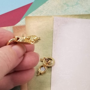 May include: Close-up of two gold-colored jewelry clasps. One clasp is held in a hand, while the other is on a light-colored surface. The clasps have intricate detailing and a magnetic closure. The background features colorful paper.
